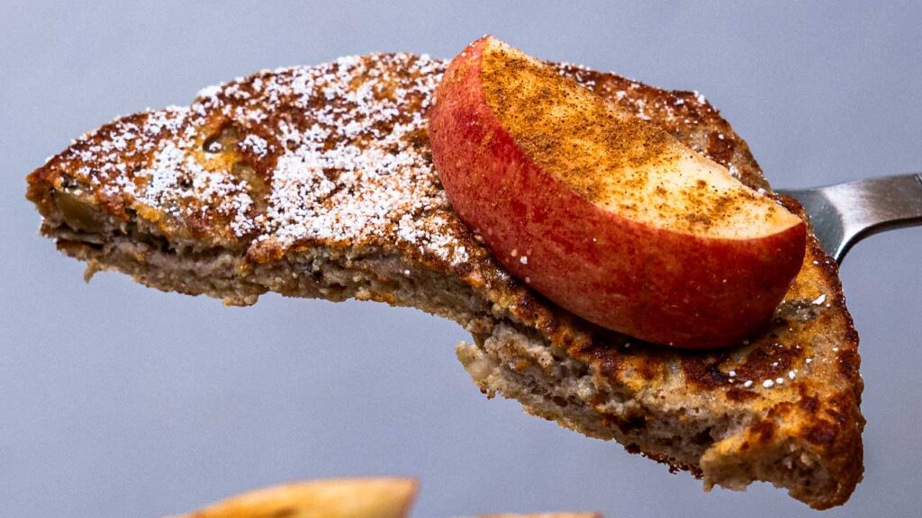 Fork lifting apple cinnamon protein french toast with an apple slice and powdered sugar