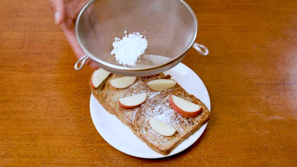 Sifting powdered sugar over apple cinnamon protein french toast topped with apple slices