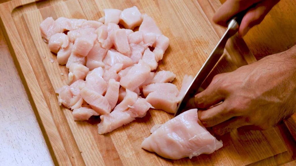 Cubing raw chicken breast on a cutting board for a high-protein pasta recipe