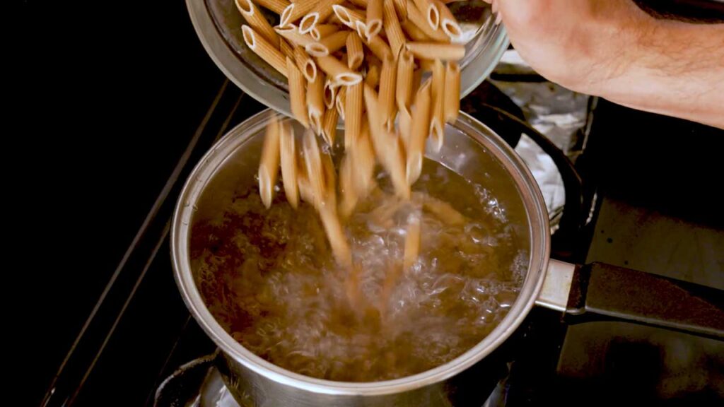 Adding whole wheat penne to boiling water for a high-protein chicken alfredo recipe