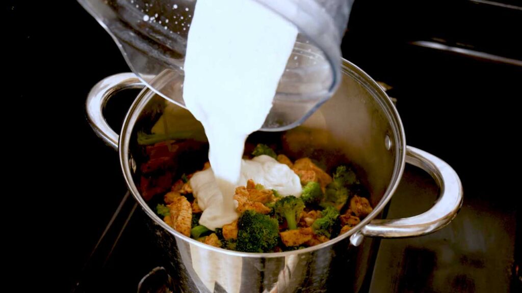 Pouring the blended cottage cheese alfredo sauce over chicken and broccoli in a pot