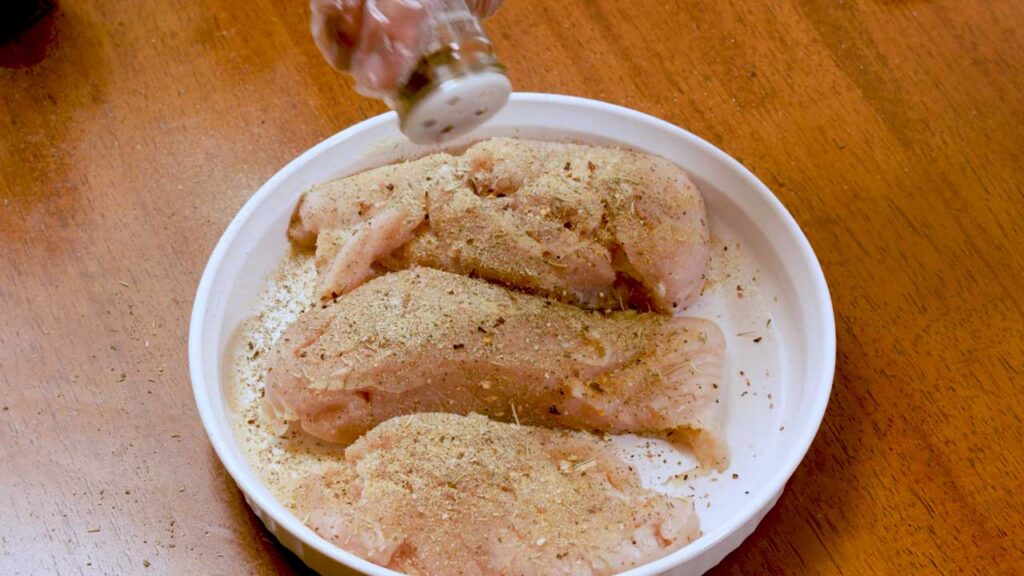 Seasoning raw chicken breasts with spices in a white dish before cooking