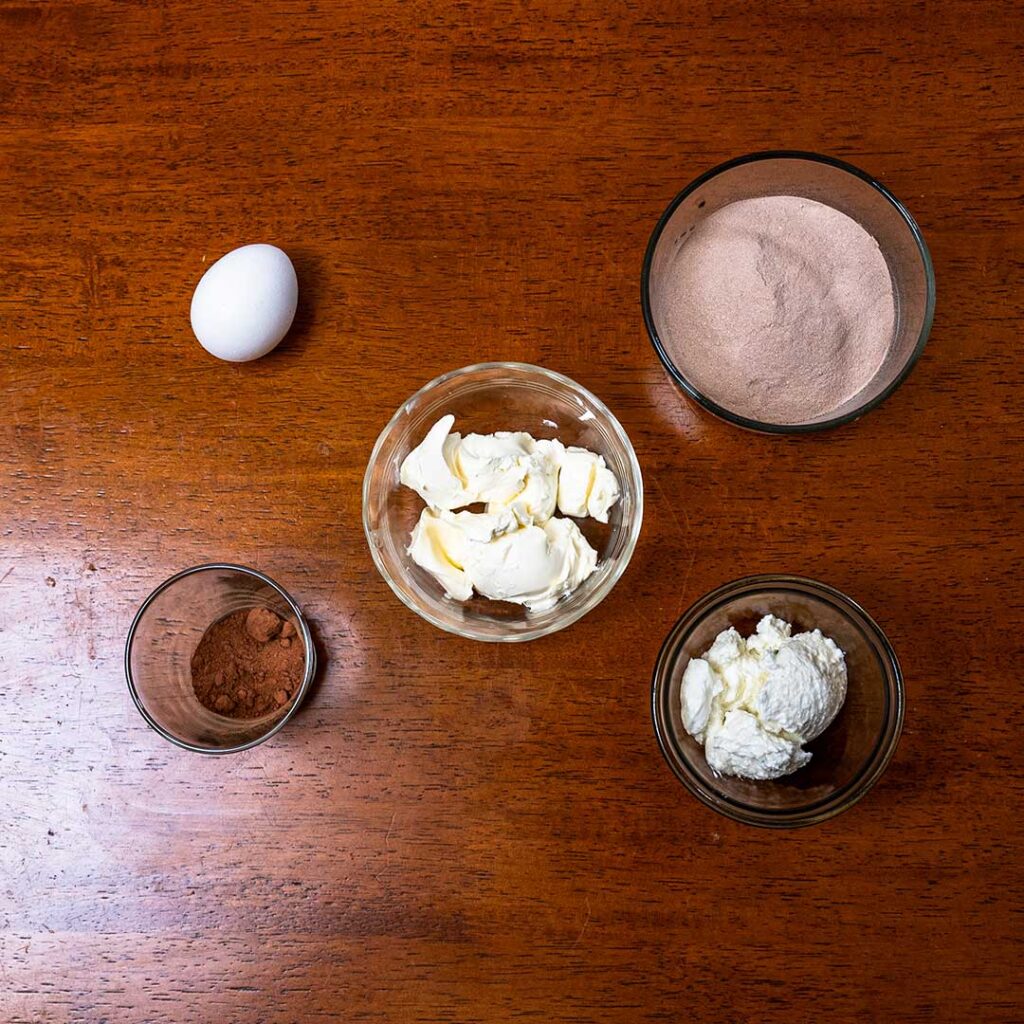 Chocolate protein cheesecake ingredients, cream cheese, Greek yogurt, cocoa powder, and an egg