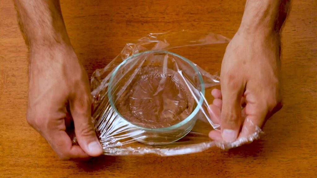 Covering the cooked chocolate protein cheesecake with plastic wrap to chill