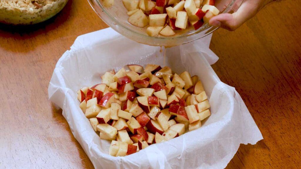 Pouring spiced apple chunks into a parchment-lined baking pan